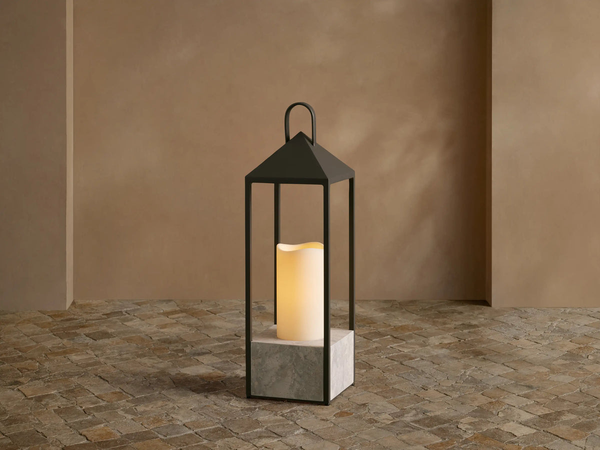 Helios Outdoor Lantern with dark bronze frame and grey marble base, featuring a glowing flameless candle on a tiled floor.