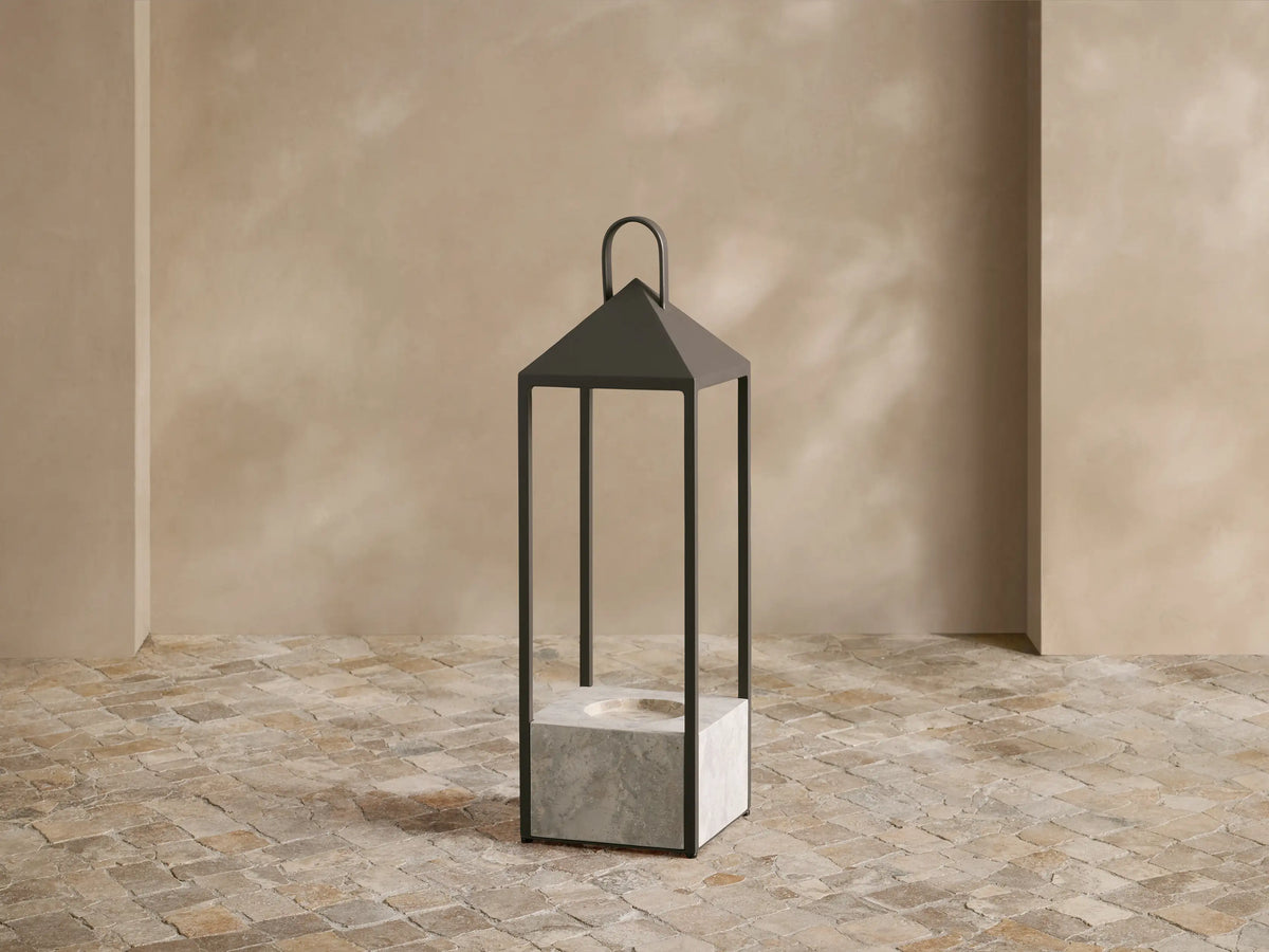 Helios Outdoor Lantern with dark bronze frame and grey marble base, featuring a glowing flameless candle on a tiled floor.