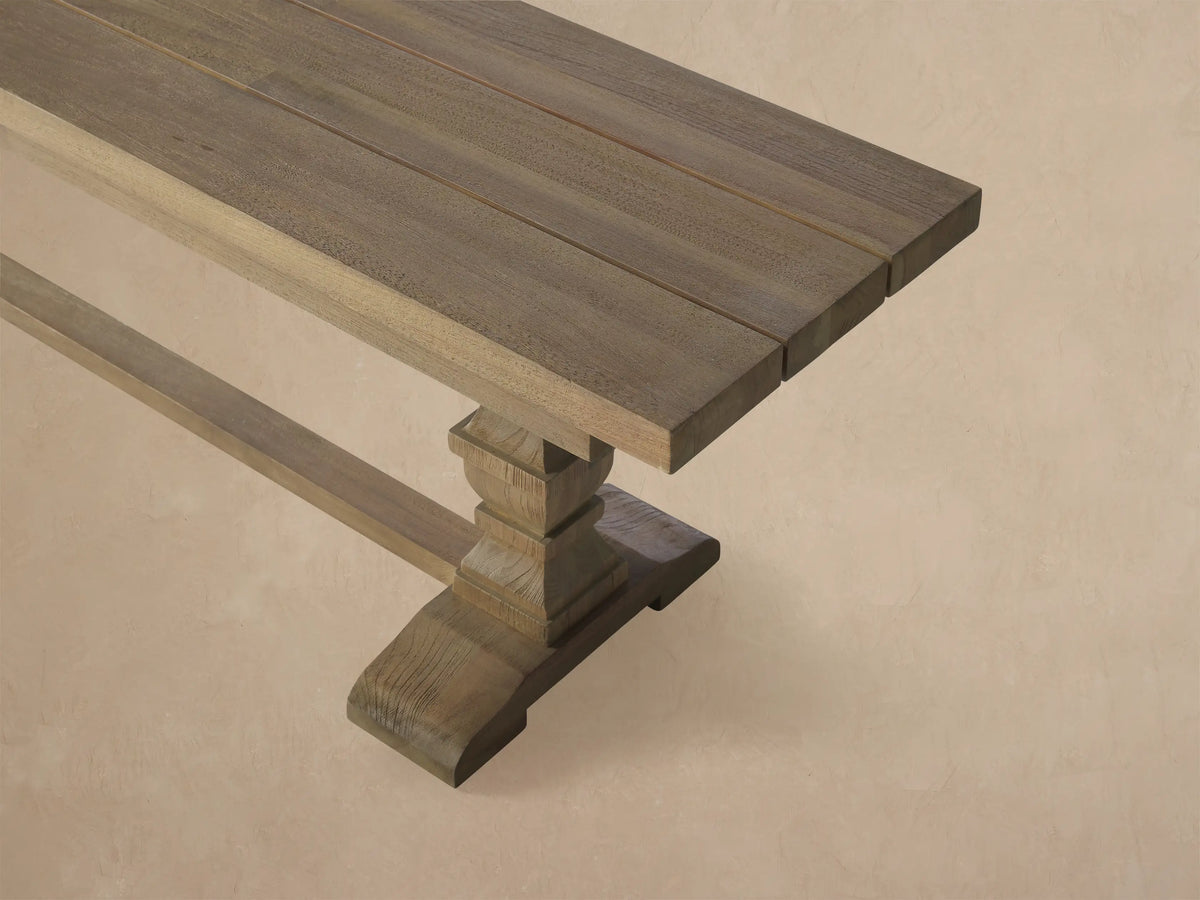 Long Hamptons Outdoor Dining Bench, light grey weathered teak, with two pedestal legs, against a wood shingle wall.
