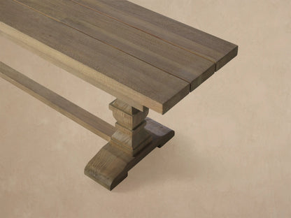 Long Hamptons Outdoor Dining Bench, light grey weathered teak, with two pedestal legs, against a wood shingle wall.