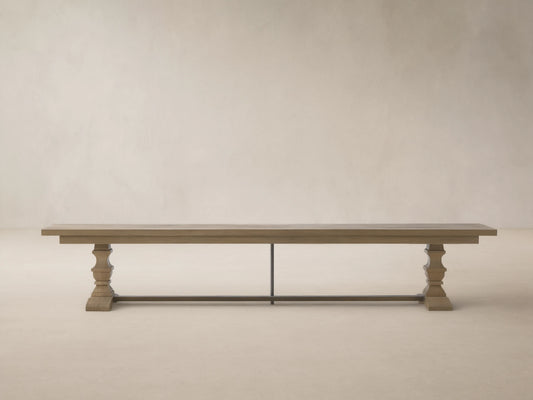 Long Hamptons Outdoor Dining Bench, light grey weathered teak, with two pedestal legs, against a wood shingle wall.