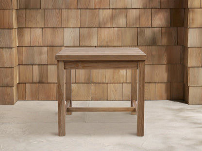 Square Hamptons Outdoor End Table, light brown natural teak wood, centered against a wood shingle wall on a light grey floor.