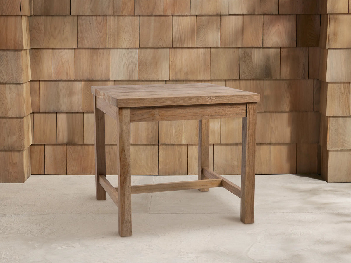 Square Hamptons Outdoor End Table, light brown natural teak wood, centered against a wood shingle wall on a light grey floor.