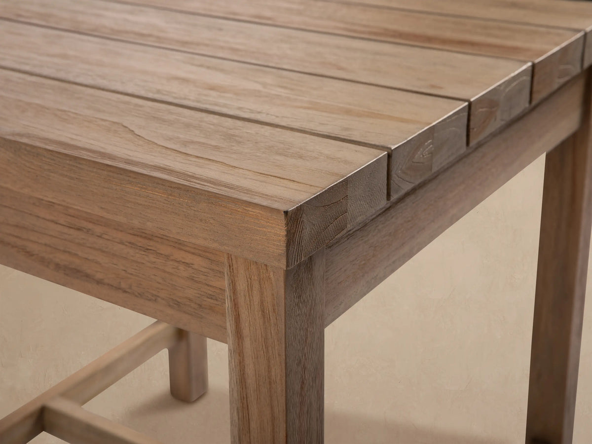 Hamptons Outdoor End Table, light natural teak wood, square slatted top, sturdy legs, on a light patio against shingle wall.