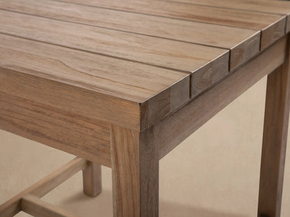 Hamptons Outdoor End Table, light natural teak wood, square slatted top, sturdy legs, on a light patio against shingle wall.