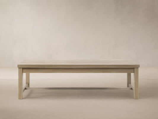 Front view of a rectangular Hamptons Outdoor Coffee Table made of light weathered teak, centered on a gray patio with a wood shingle wall.