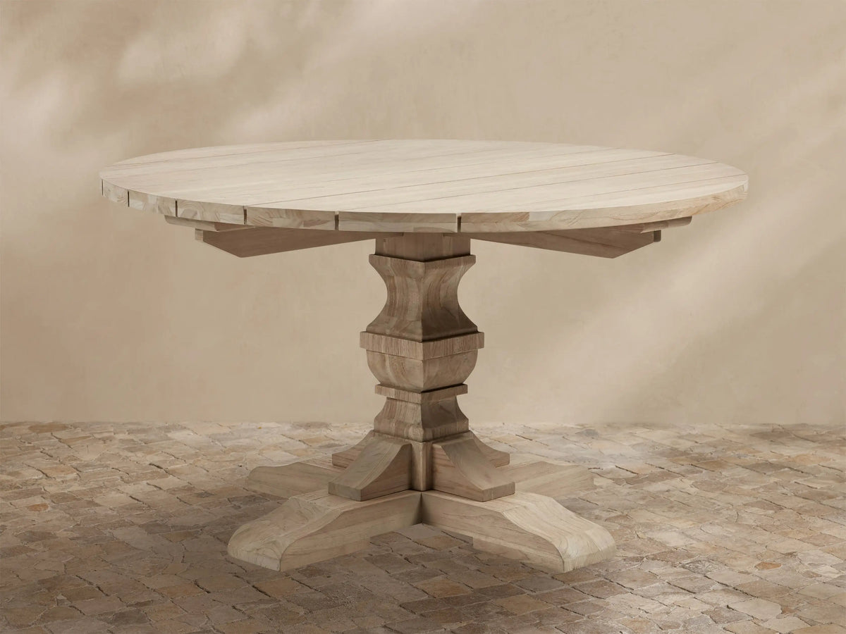 Light teak Hamptons Outdoor Round Dining Table, with carved pedestal base & splayed feet, on a patterned patio.