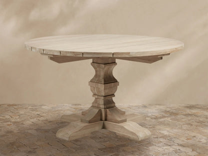 Light teak Hamptons Outdoor Round Dining Table, with carved pedestal base & splayed feet, on a patterned patio.
