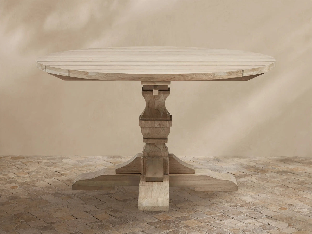 Light teak Hamptons Outdoor Round Dining Table, with carved pedestal base & splayed feet, on a patterned patio.