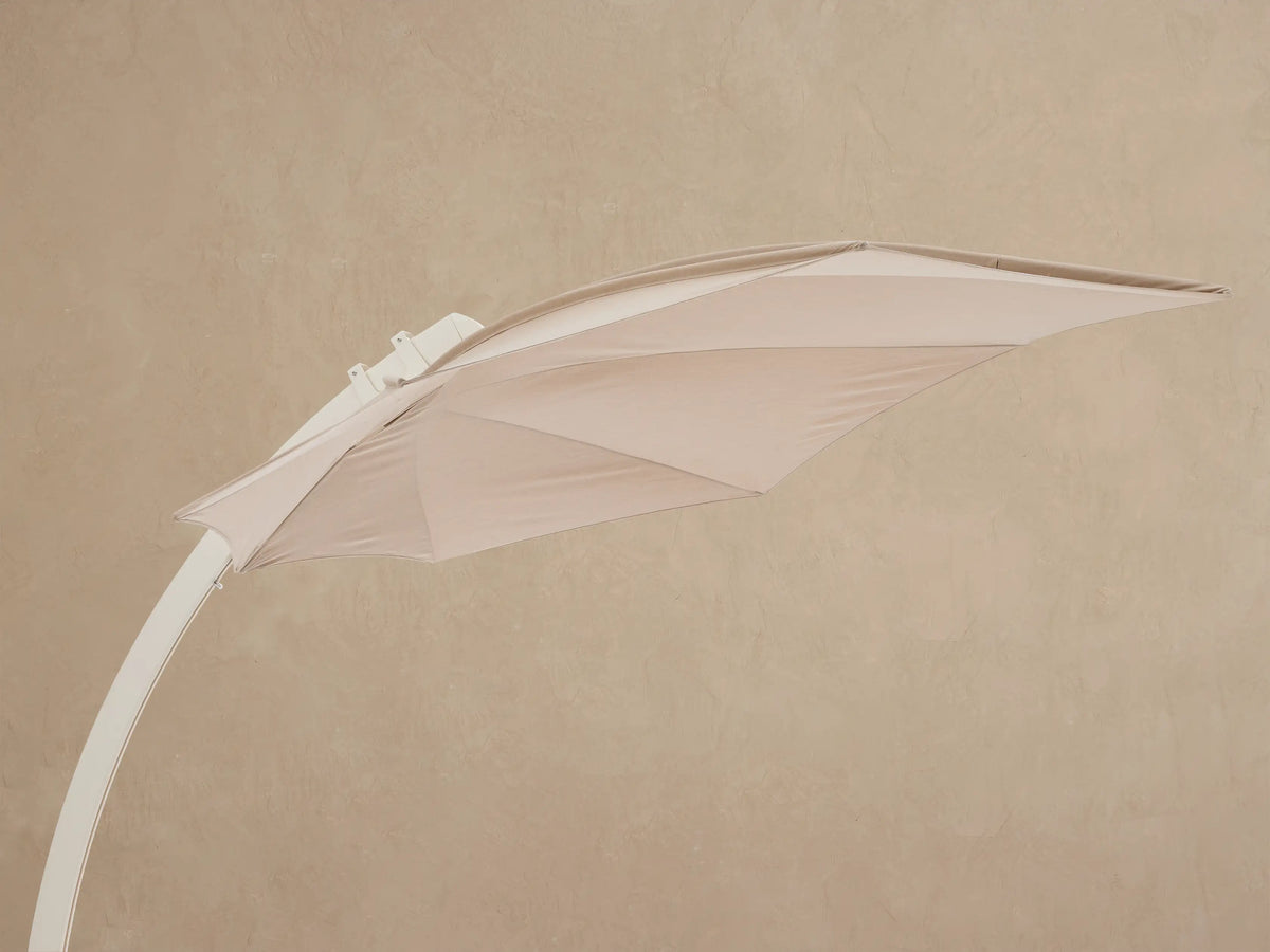 Close-up of Icarus Outdoor Umbrella in sand. Beige canopy, cream pole with silver handle, white base on slatted wood surface.