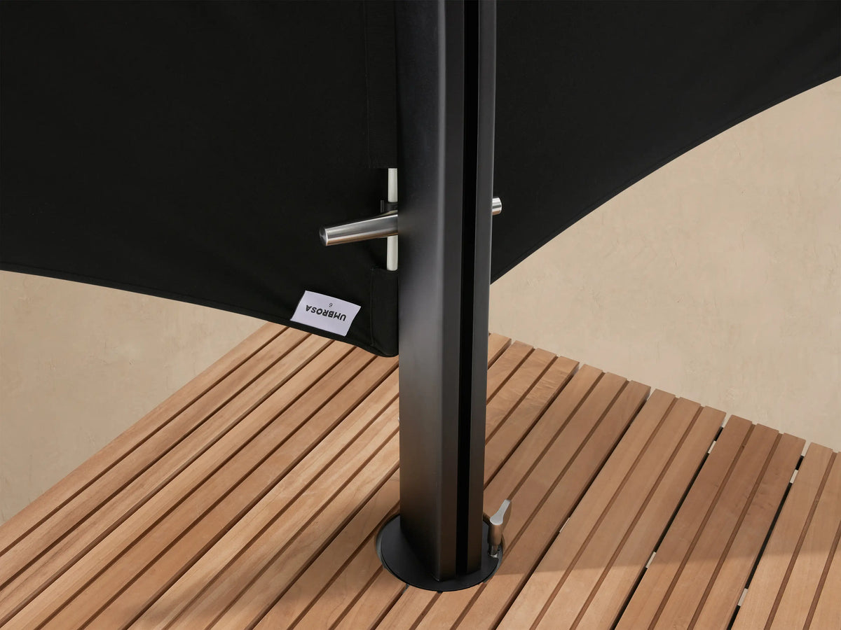 Black Icarus Outdoor Umbrella, folded and angled sharply upwards, on a slatted wood-topped wheeled base, against a light wall.
