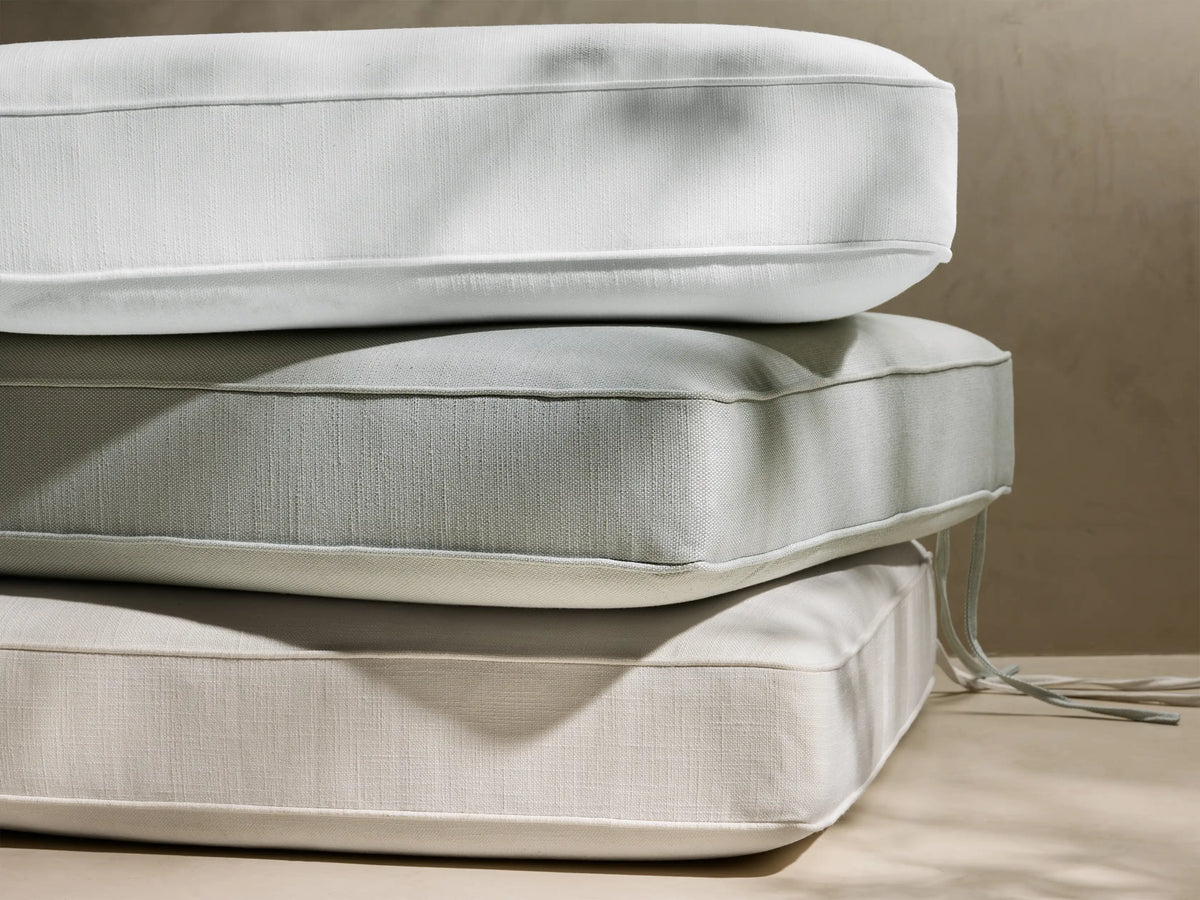 Stacked Ilia outdoor water-repellent cushions: white, light gray, beige. Piped edges, textured fabric, tie strings visible. Dappled light.