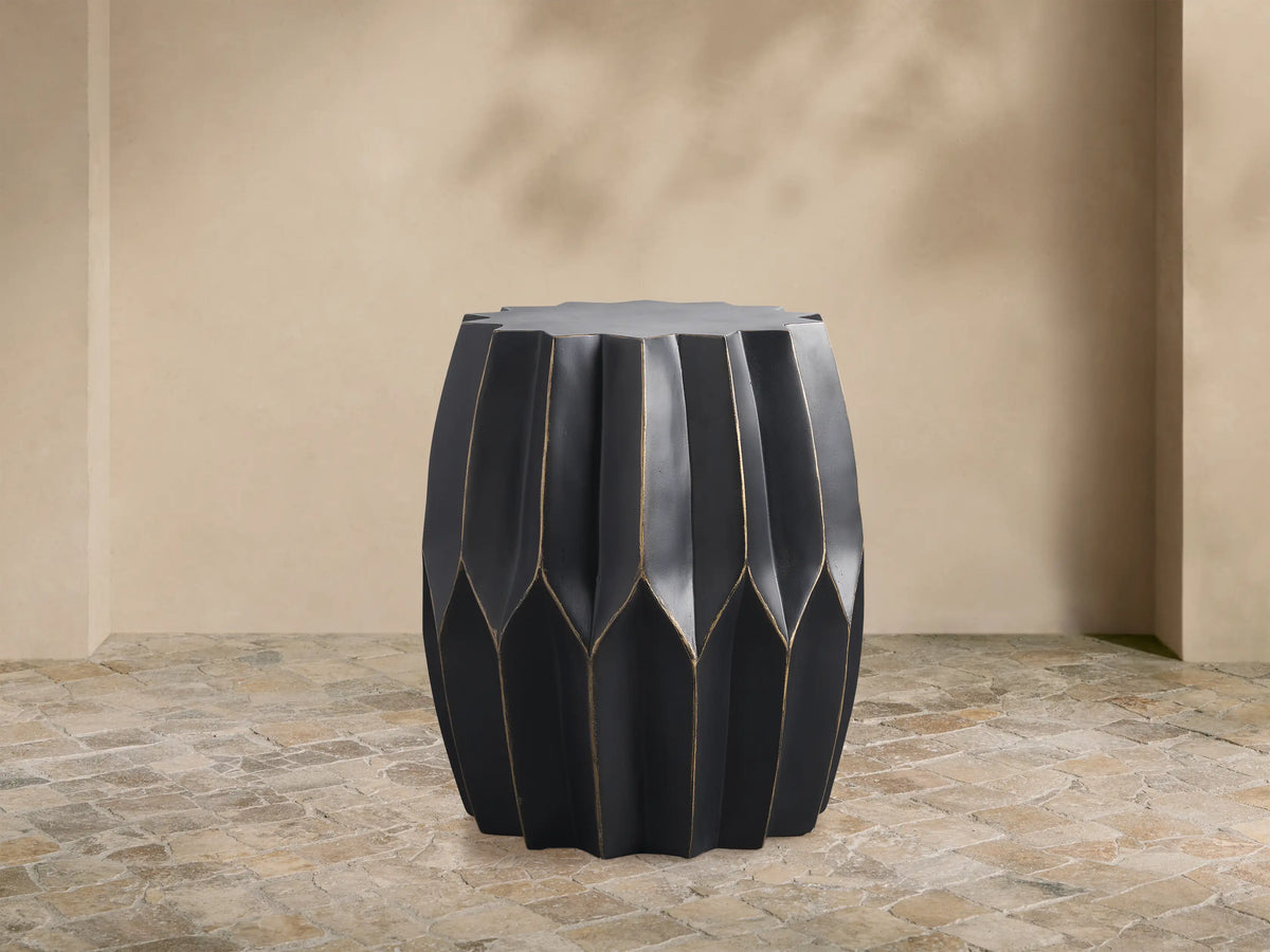 Indio Outdoor End Table, dark gray with gold geometric facets, standing on a stone patio against a tan wall.