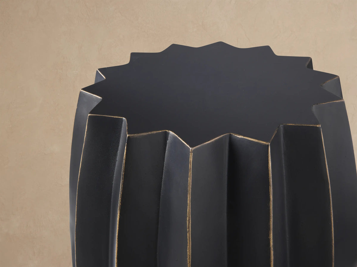 Indio Outdoor End Table, dark gray with gold geometric facets, standing on a stone patio against a tan wall.