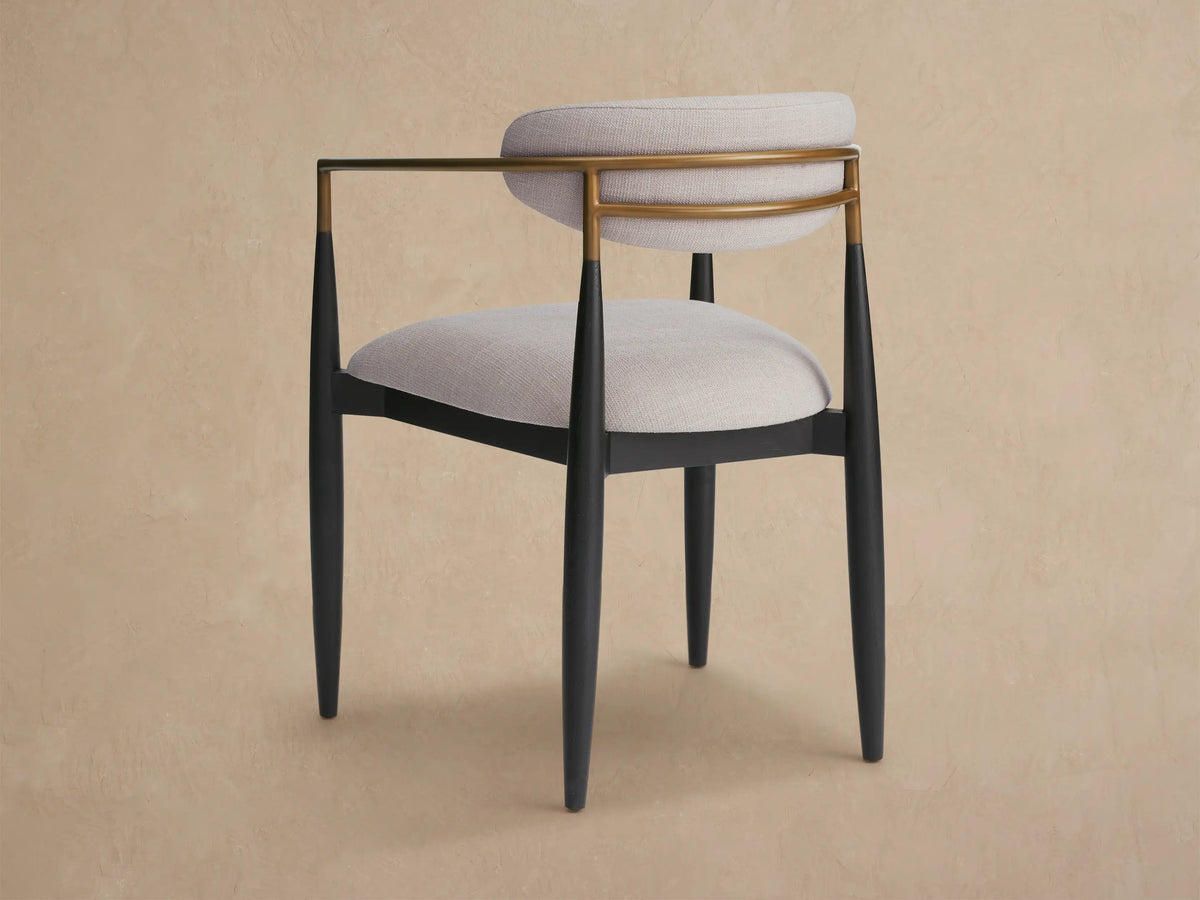 Angled close-up of Jagger Outdoor Dining Arm Chair with curved light beige cushion, brass-toned frame, and dark leg.