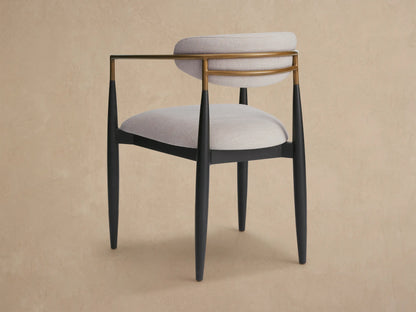 Angled close-up of Jagger Outdoor Dining Arm Chair with curved light beige cushion, brass-toned frame, and dark leg.