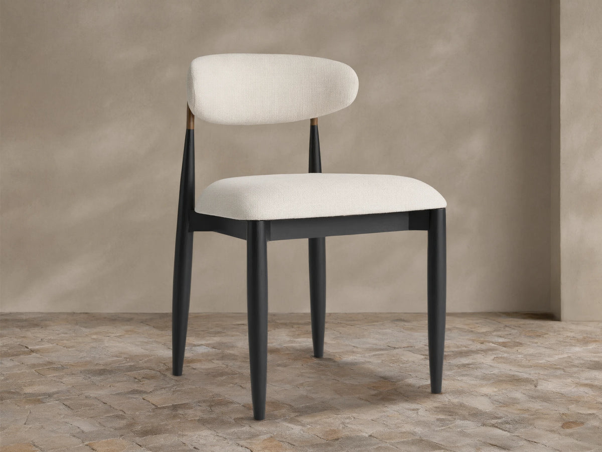 Jagger Outdoor Dining Side Chair: sleek black wood frame, curved off-white upholstered seat and back, brass accent, on stone floor.
