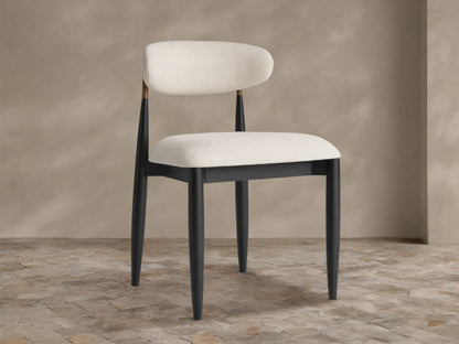 Jagger Outdoor Dining Side Chair: sleek black wood frame, curved off-white upholstered seat and back, brass accent, on stone floor.