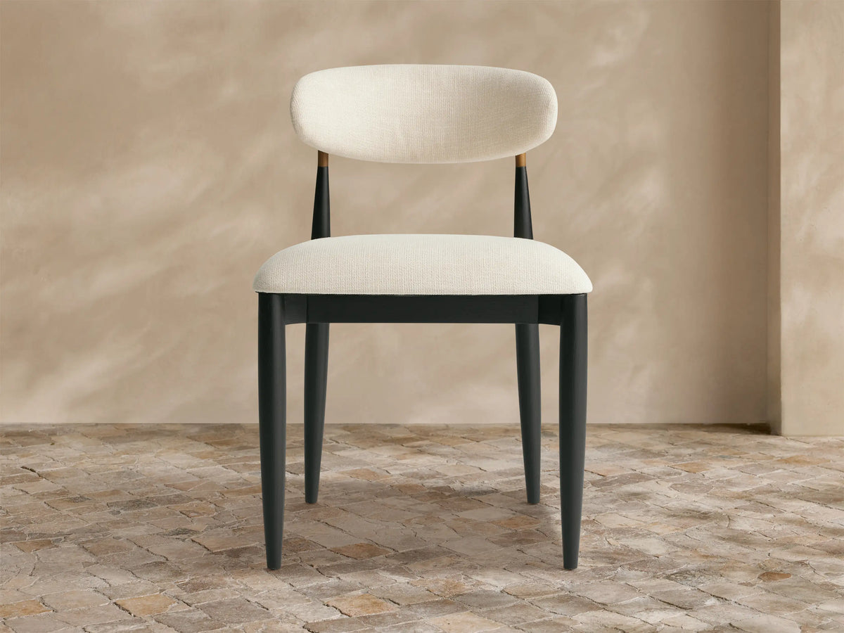 Jagger Outdoor Dining Side Chair: sleek black wood frame, curved off-white upholstered seat and back, brass accent, on stone floor.
