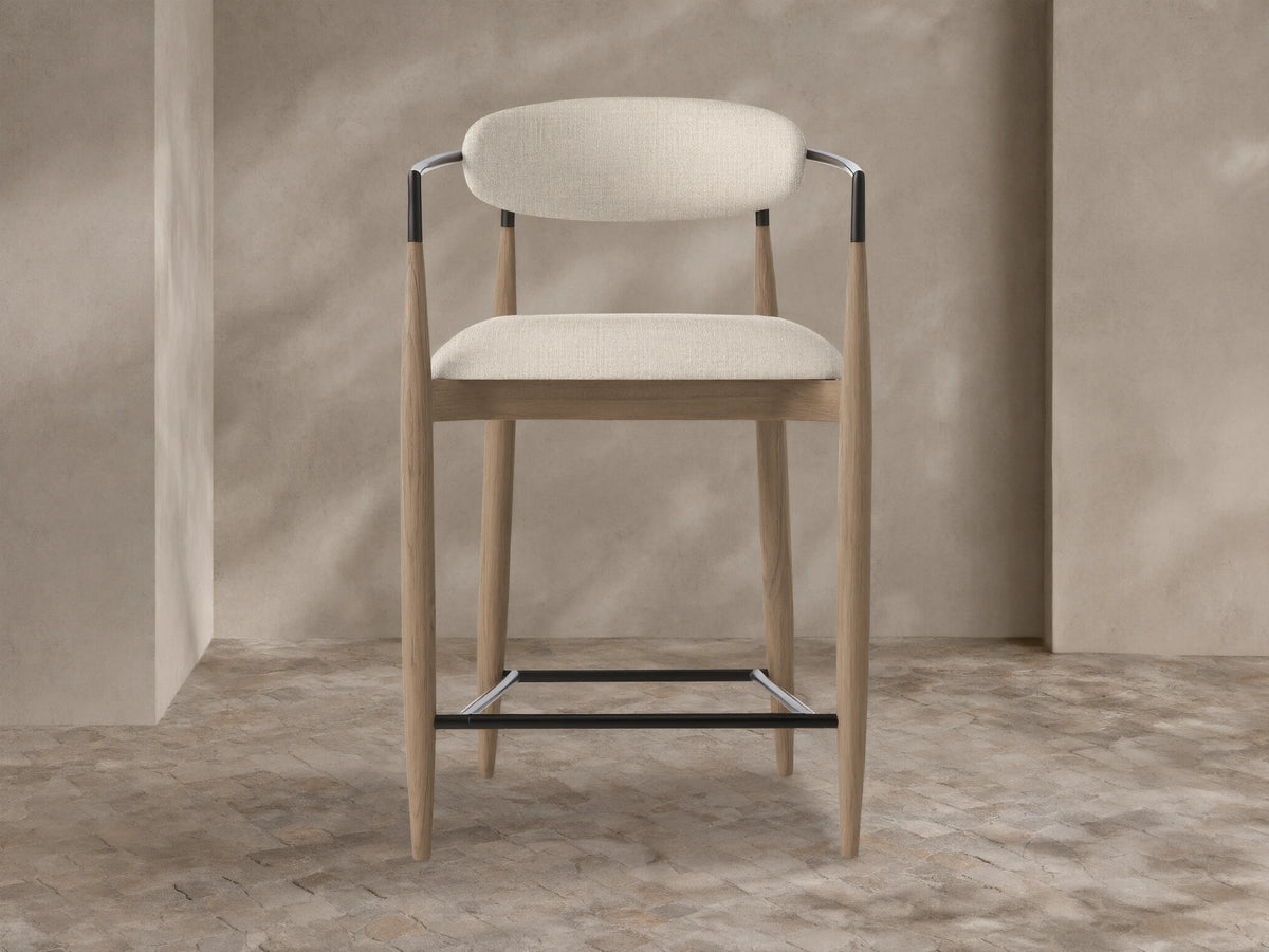 Jagger Outdoor Counter Stool with natural teak frame, light beige upholstered seat & back, black metal armrests & footrest.
