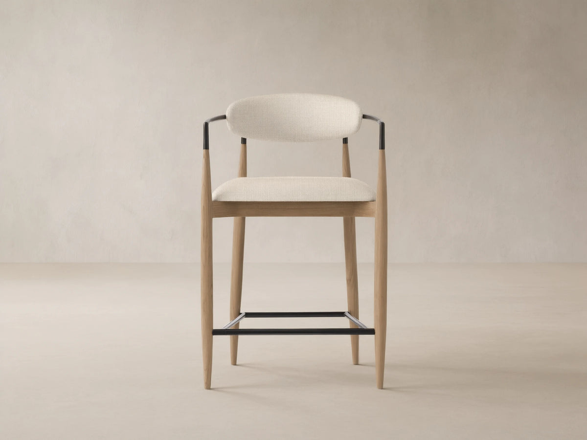 Jagger Outdoor Counter Stool with natural teak frame, light beige upholstered seat & back, black metal armrests & footrest.