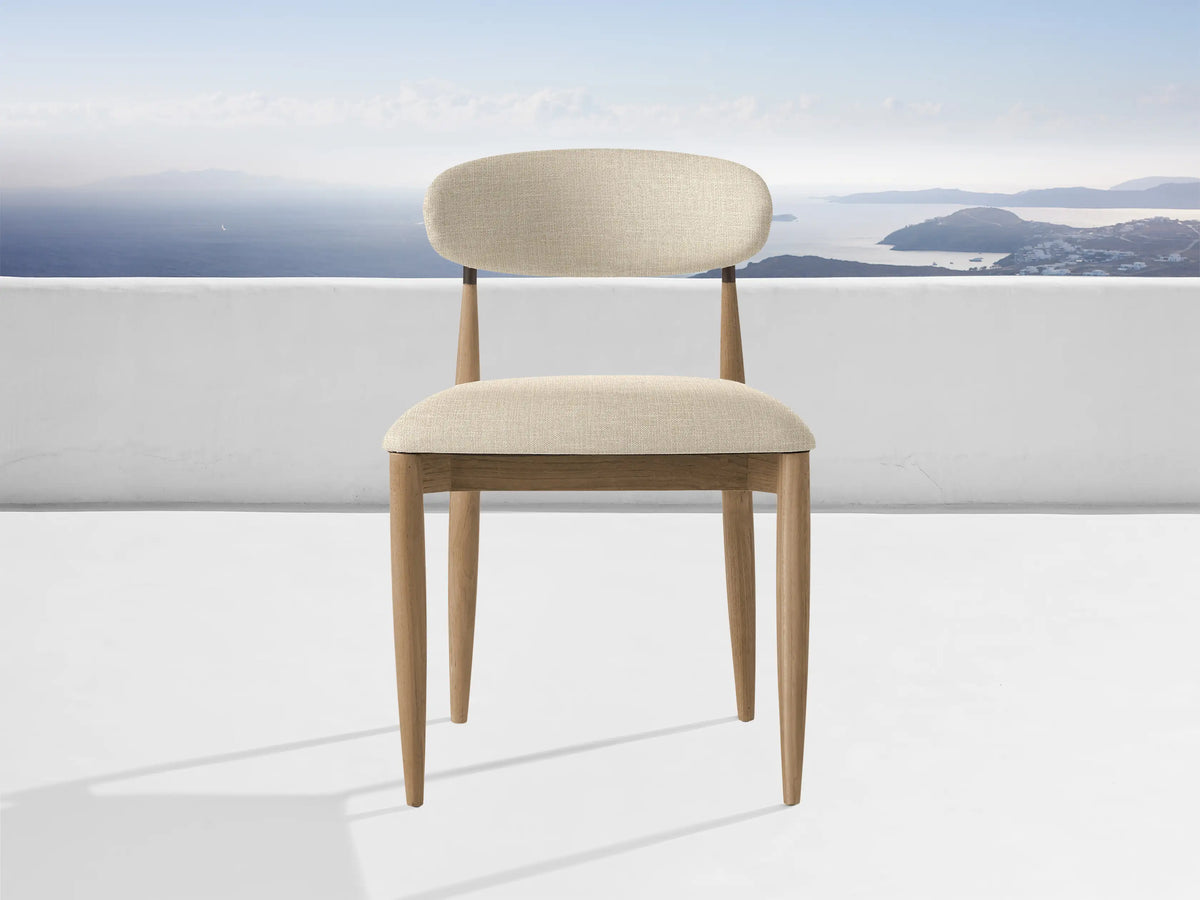Jagger Outdoor Dining Side Chair, natural teak frame, light beige cushions, on a white patio overlooking a blue sea & islands.