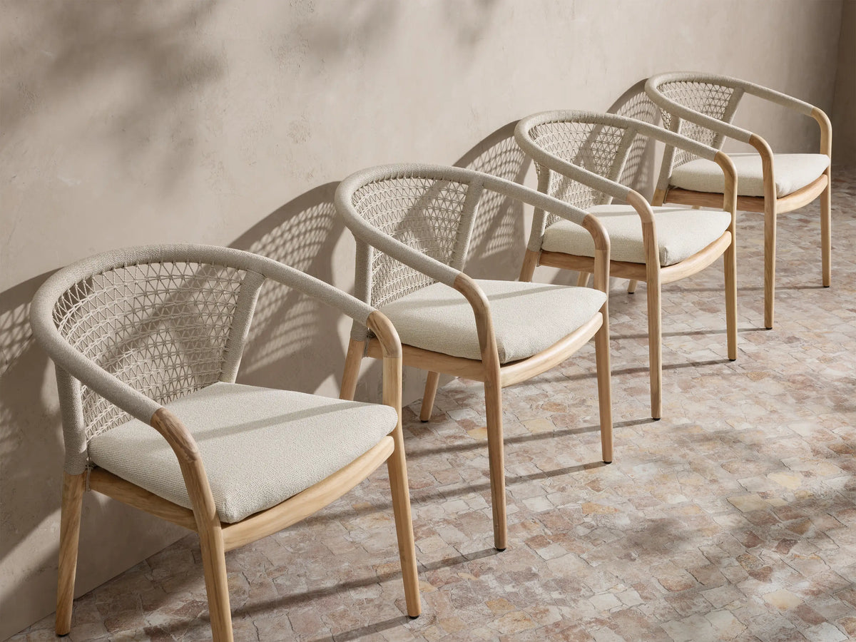 Four Kaia Outdoor Dining Chair with Cushion chairs with natural teak frames, light beige woven backs, and light beige cushions.