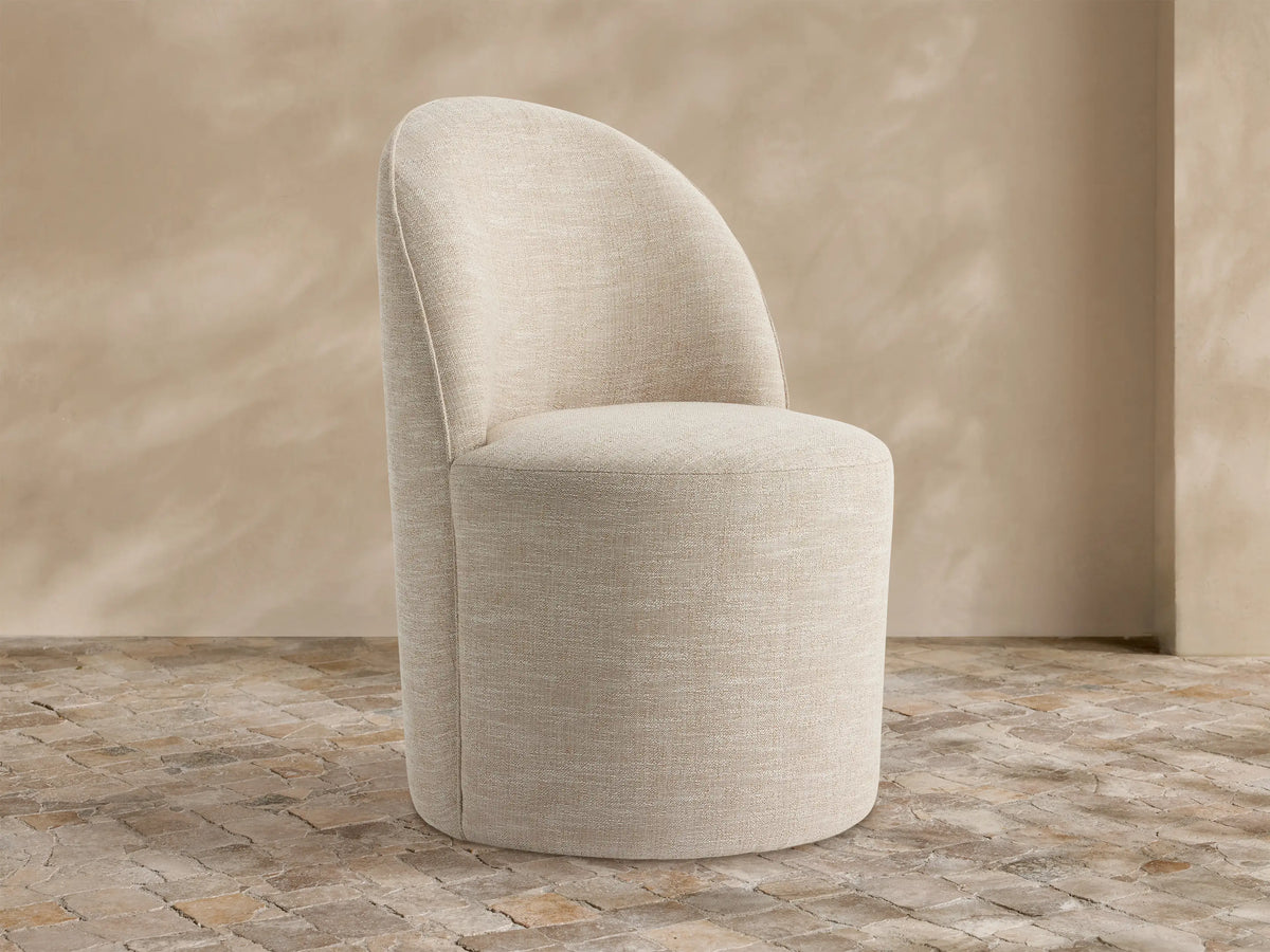 Kora Outdoor Dining Chair, light beige textured fabric, curved back, round seat, on rustic stone tiles against a textured tan wall.