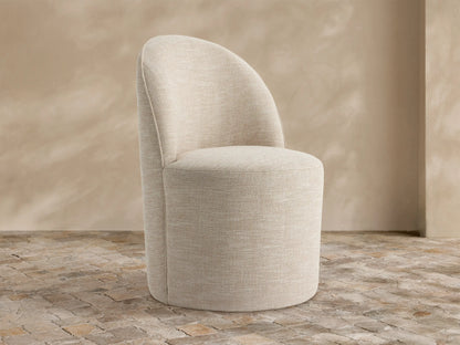 Kora Outdoor Dining Chair, light beige textured fabric, curved back, round seat, on rustic stone tiles against a textured tan wall.