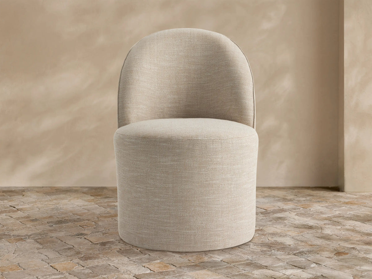 Kora Outdoor Dining Chair, light beige textured fabric, curved back, round seat, on rustic stone tiles against a textured tan wall.