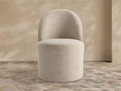 Kora Outdoor Dining Chair, light beige textured fabric, curved back, round seat, on rustic stone tiles against a textured tan wall.