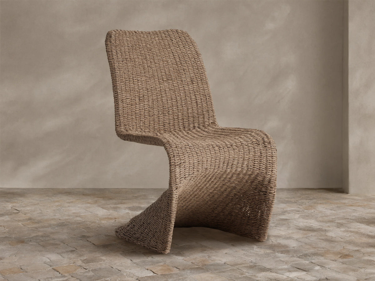 Leon Outdoor Dining Chair, curved, natural brown woven resin wicker, against a textured beige wall on light stone tiles.