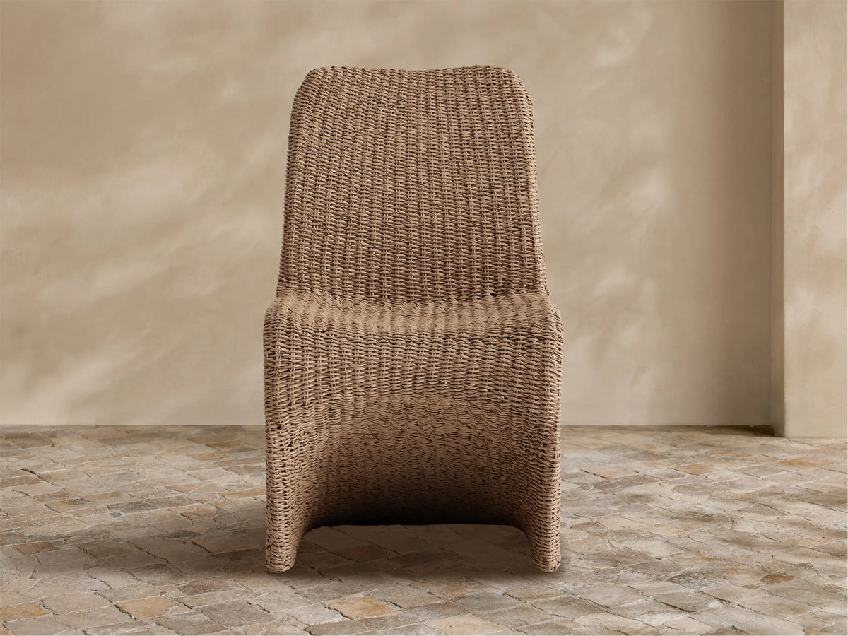 Leon Outdoor Dining Chair, curved, natural brown woven resin wicker, against a textured beige wall on light stone tiles.