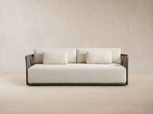 Lisbon Outdoor Sofa with light cream cushions and a dark brown woven rope frame on a tiled patio against a beige wall.