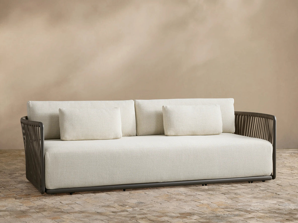 Lisbon Outdoor Sofa with light cream cushions and a dark brown woven rope frame on a tiled patio against a beige wall.