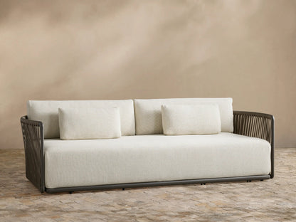 Lisbon Outdoor Sofa with light cream cushions and a dark brown woven rope frame on a tiled patio against a beige wall.