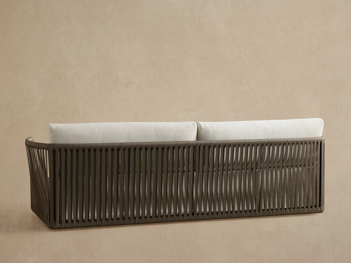 Angled view of Lisbon Outdoor Sofa. Cream fabric seat and back cushions, lumbar pillow, dark frame with brown woven rope arm.