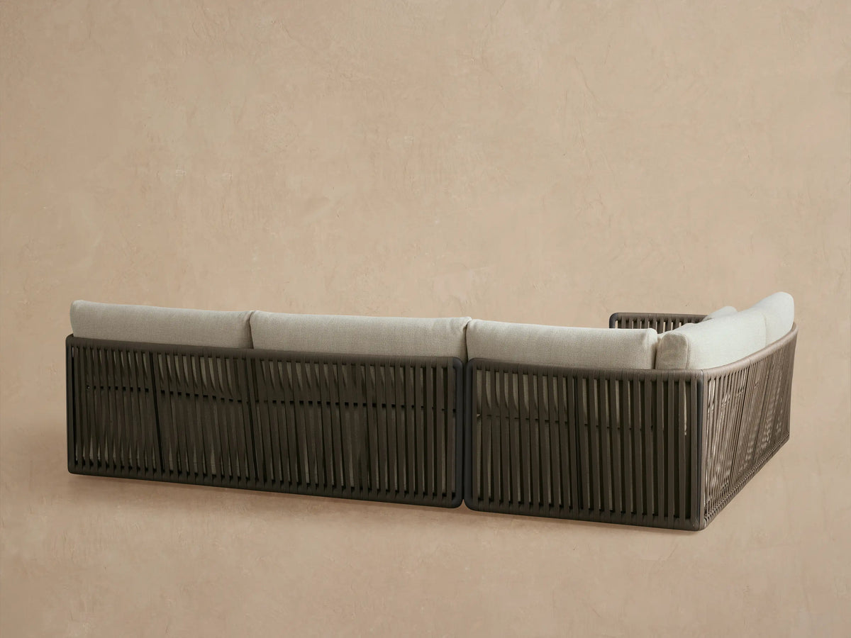 Close-up of Lisbon Outdoor Sectional showing dark gray woven rope frame, light beige cushions in soft focus behind.