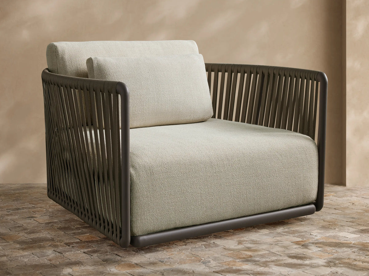 Lisbon Outdoor Swivel Chair with dark gray woven frame, light beige cushions, angled on a paved patio against a beige wall.