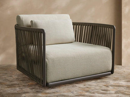 Lisbon Outdoor Swivel Chair with dark gray woven frame, light beige cushions, angled on a paved patio against a beige wall.