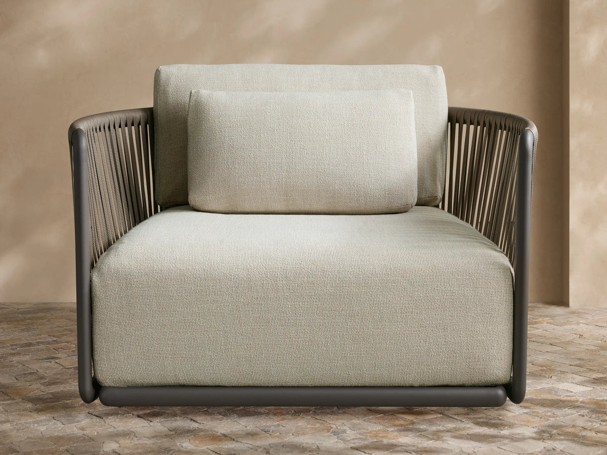 Lisbon Outdoor Swivel Chair with dark gray woven frame, light beige cushions, angled on a paved patio against a beige wall.