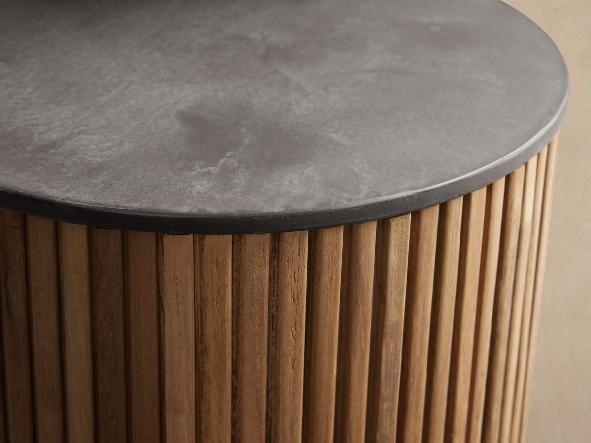 Pompeii Outdoor End Table, cylindrical, natural teak wood slatted base, dark grey concrete top, on stone patio.