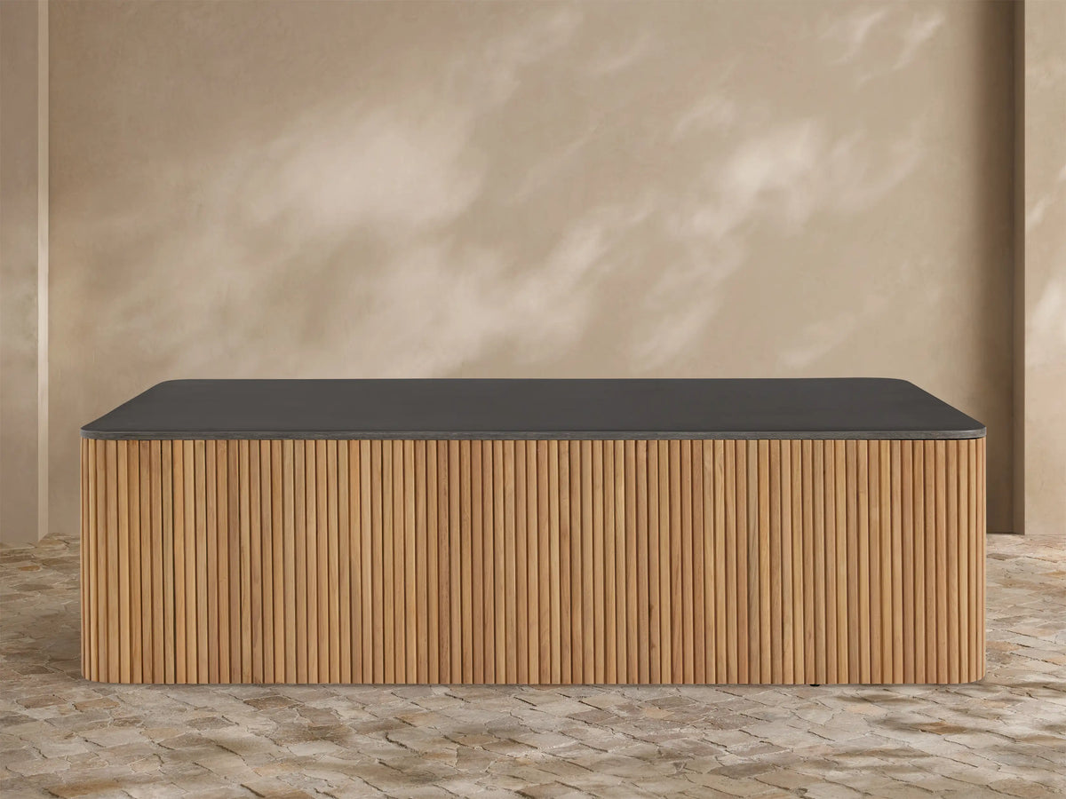 Pompeii Outdoor Coffee Table with a dark gray concrete top and slatted natural teak wood base, set against a beige wall.
