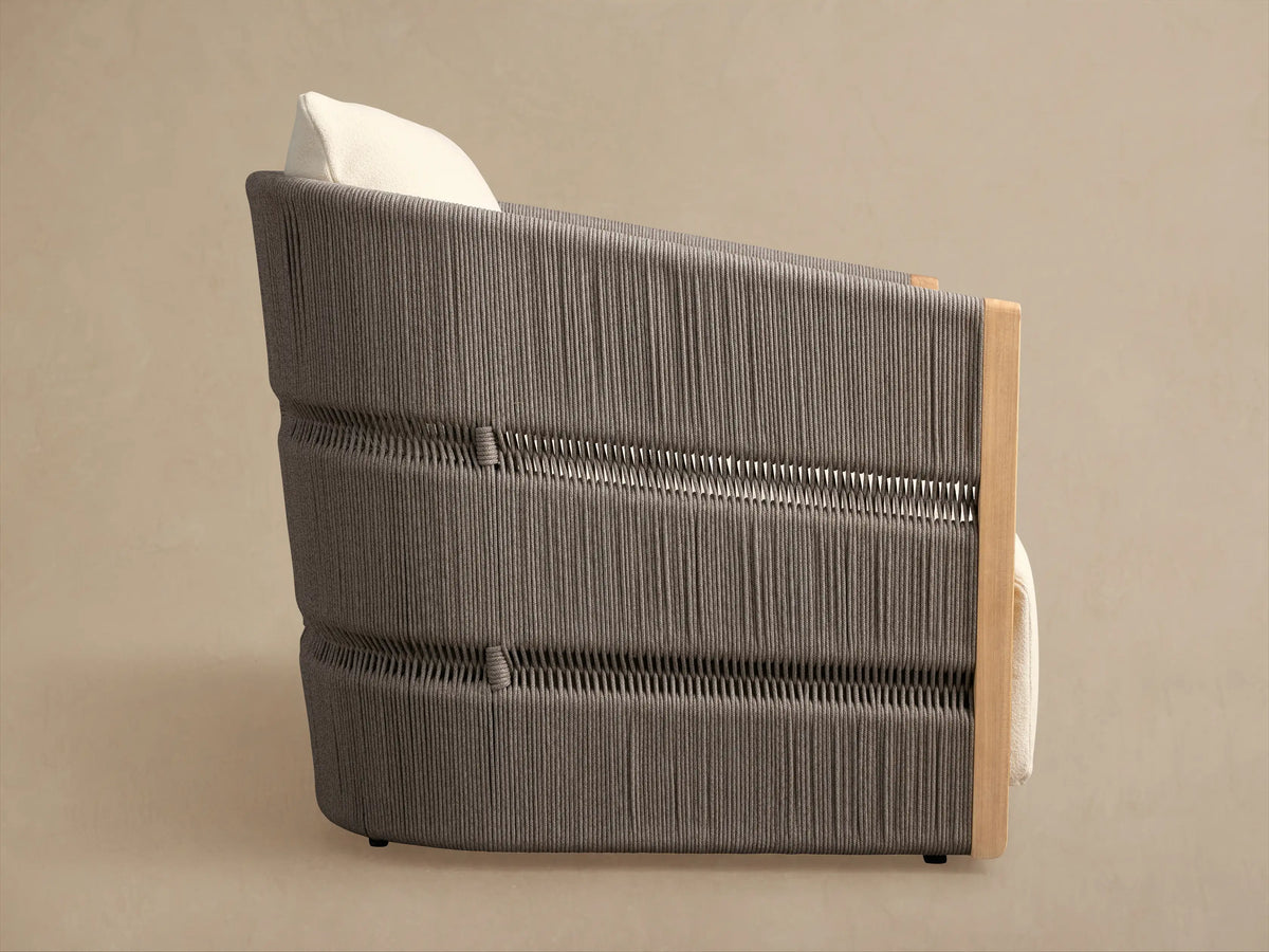 Side view of Manadala Outdoor Swivel Chair. Dark gray handwoven rope texture, light teak wood frame, and ivory cushion.