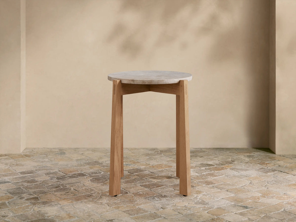 Mari Outdoor End Table with round, light gray marble top and light teak wood legs on a rustic tiled floor.