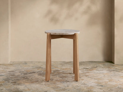 Mari Outdoor End Table with round, light gray marble top and light teak wood legs on a rustic tiled floor.