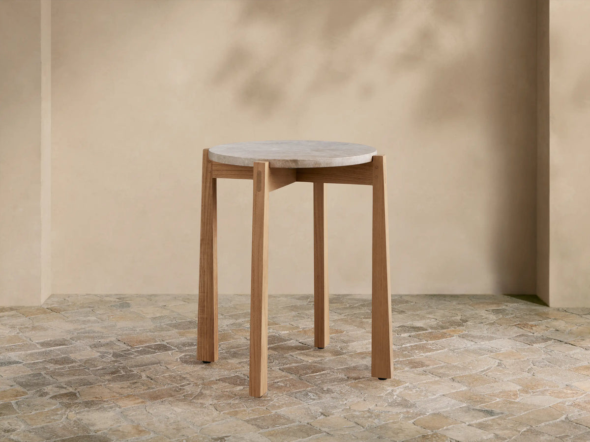 Mari Outdoor End Table with round, light gray marble top and light teak wood legs on a rustic tiled floor.