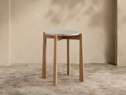 Mari Outdoor End Table with round, light gray marble top and light teak wood legs on a rustic tiled floor.