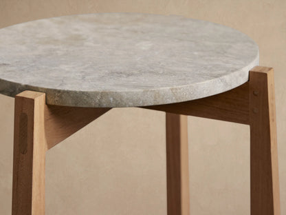 Mari Outdoor End Table with round grey marble top and light teak wood legs, on a stone patio against a tan wall with leaf shadows.