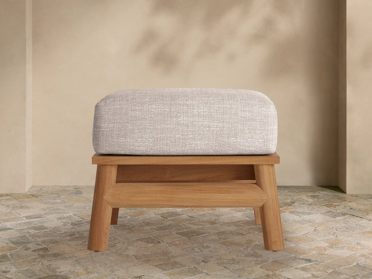 Maxime Outdoor Ottoman with a light gray textured cushion on a natural teak wood base, set on a stone patio with a light beige wall.
